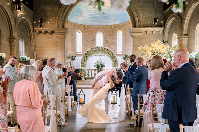 A bride in a white wedding dress and a groom in a pink suit dip kissing as the come back up the aisle after their wedding vows surrounded by their guests at Sneaton Castle in Whitby, Yorkshire