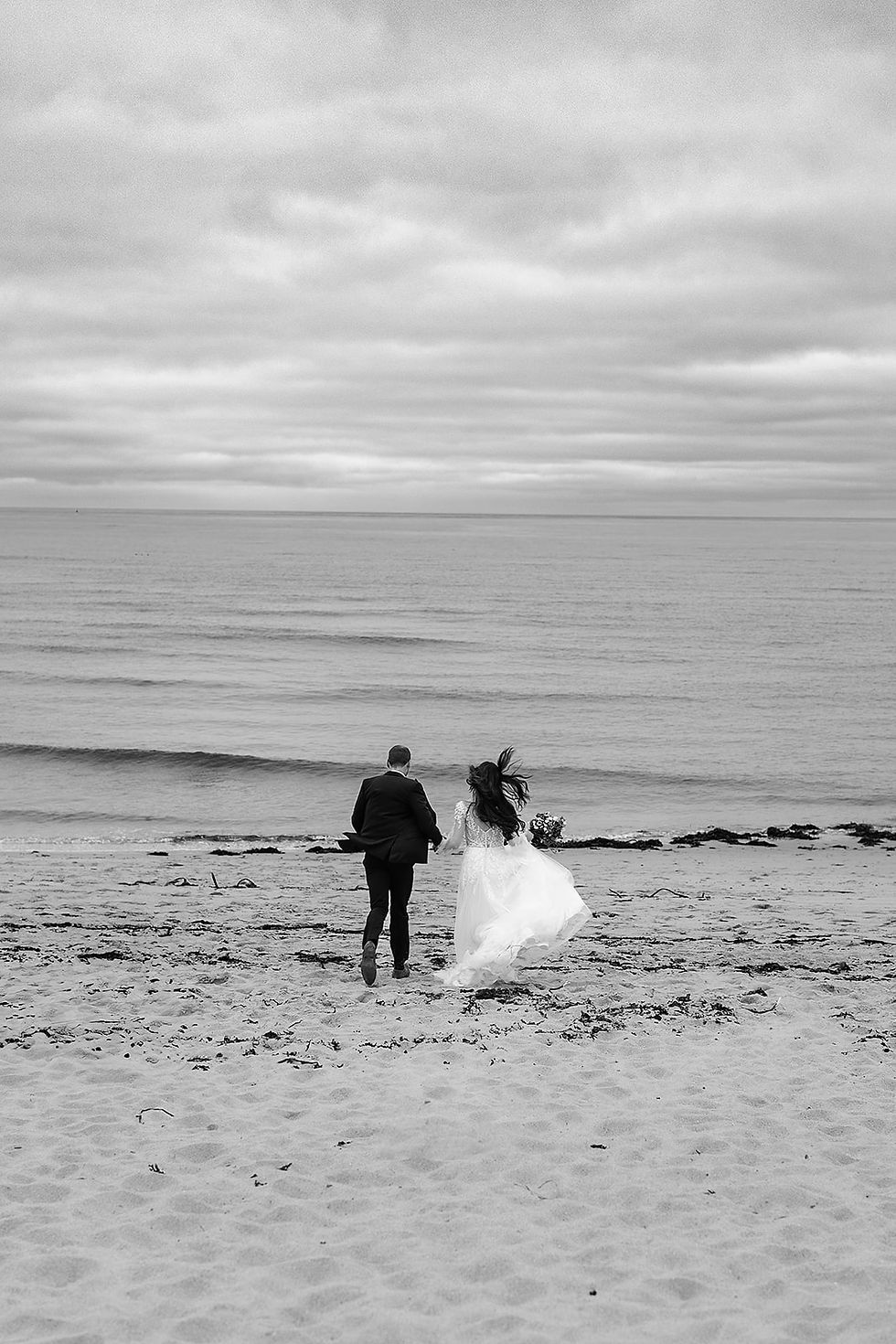 A couple in white wedding attire running towards the beach