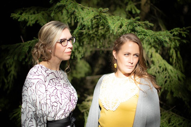 Elina Silvo ja Anna Karvosenoja kuvattuna kuusen edessä, taustalla näkyvät vain kuusen okset ja naiset katsovat poispäin kamerasta oikealle, ikään kuin toiseen kameraan.