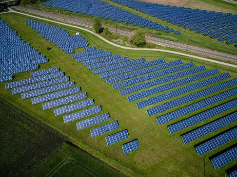 Aerial view of a solar farm with rows of blue solar panels on green grass, divided by a dirt path. Overcast sky enhances the calm mood.