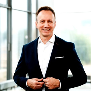 Smiling man in a navy suit adjusts his jacket in a bright, modern office. Glass walls and city view in the background.
