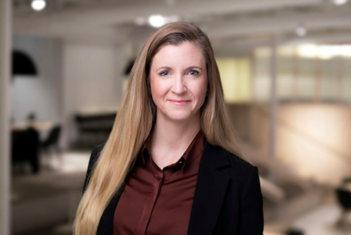 Smiling woman with long hair in a black blazer and maroon shirt indoors, blurred office background, creating a professional atmosphere.
