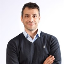 Smiling man with crossed arms in a dark sweater over a blue shirt against a plain white background, exuding confidence.