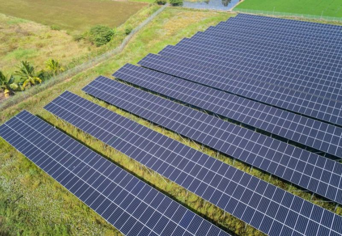 Rows of solar panels on green grass under a partly cloudy sky. Panels are dark blue-black with a grid pattern, surrounded by lush fields.