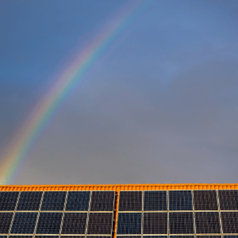 Rainbow arcs over rows of solar panels beneath a cloudy blue sky, creating a calm, hopeful scene.