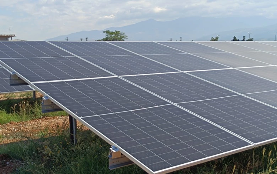Rows of solar panels in a grassy field under a cloudy sky, with distant mountains in the background, capturing sunlight efficiently.