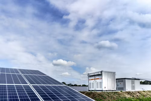 Solar panels in foreground beside white utility containers at a solar farm under a cloudy sky.