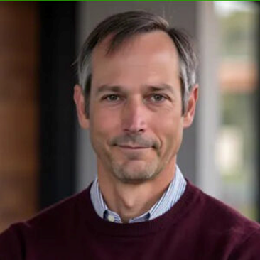 A man in a maroon sweater with a calm expression stands indoors against a blurred background, featuring soft, neutral colors.