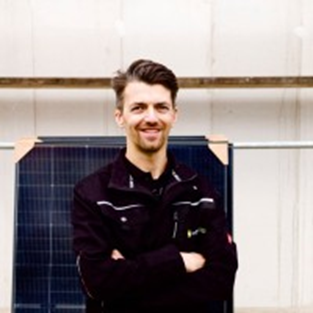 Man smiling with arms crossed in front of solar panel, wearing a dark jacket. Background shows a light structure. Bright, professional mood.