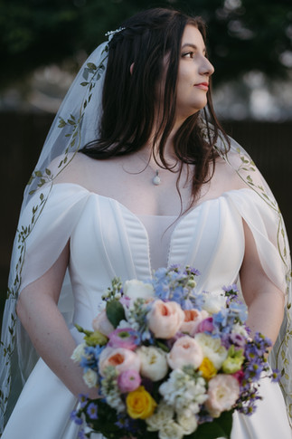 A bride in an off-the-shoulder white gown and embroidered veil holds a colorful bouquet while gazing thoughtfully to the side.