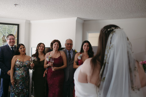 A group of smiling family members and bridesmaids react with joy as the bride, seen from behind, enters the room in her wedding dress and veil.