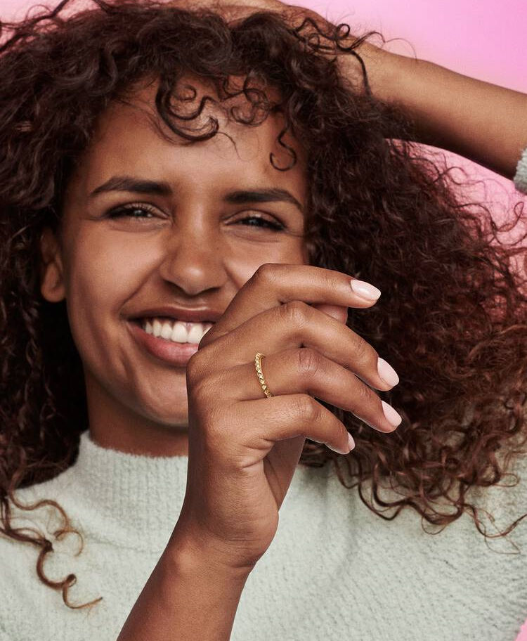 Smiling woman wears Pandora ME Yellow Gold Pyramids Ring, hand near face, cheerful expression.
