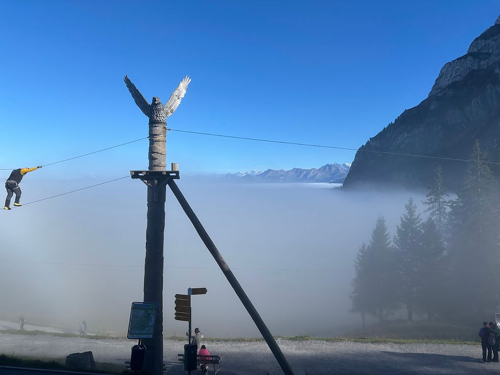 Person tightrope walking between poles with eagle sculpture, set against misty mountain backdrop. Blue sky, foggy valley, trees below.