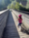 Child in pink tank top and helmet rides a balance bike on a wooden bridge, surrounded by greenery. Sunlight casts shadows on the path.