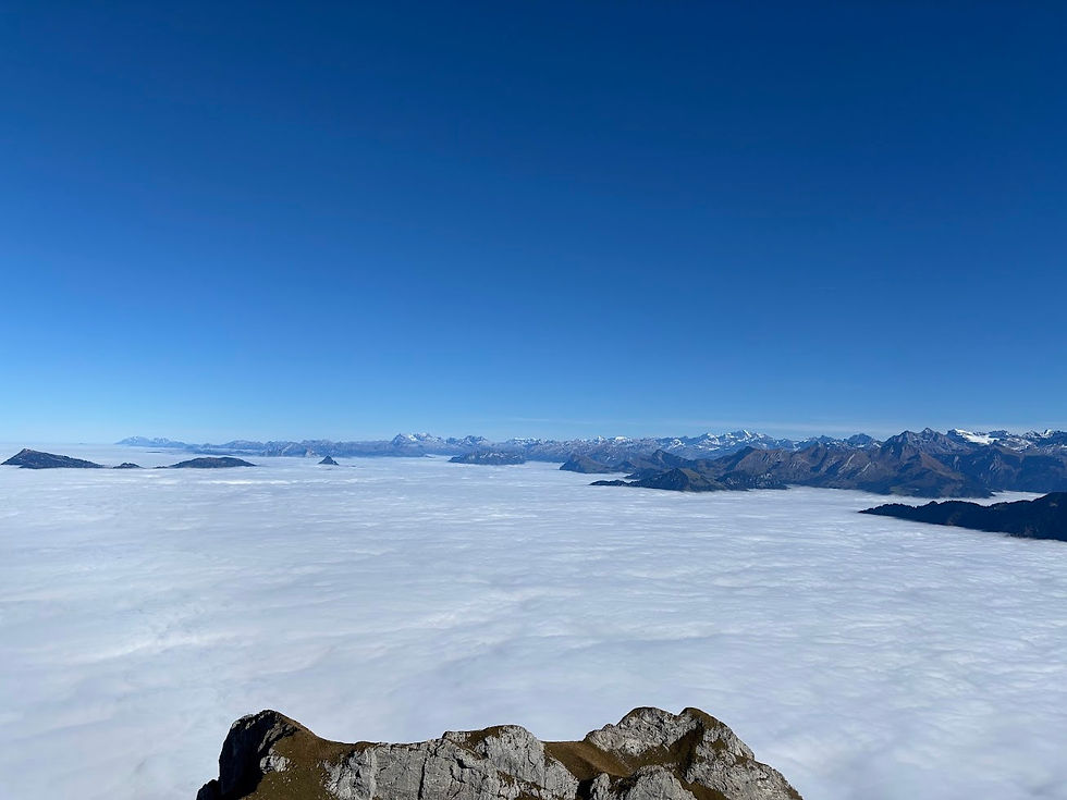 Vast mountain range under clear blue sky, above a sea of clouds. Foreground features rocky outcrop, creating a serene, majestic view.