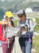 Two women in hiking gear study a map on a mountain trail. One wears a yellow cap, the other a gray cap. Clear sky and grassy hills behind.