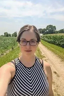 Woman wearing glasses standing on a rural dirt road beside green cornfields on a sunny day.