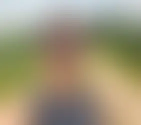 Woman wearing glasses standing on a rural dirt road beside green cornfields on a sunny day.