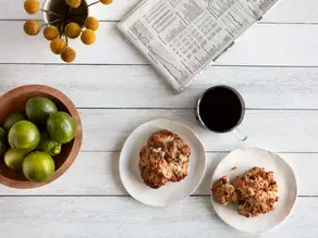 Overhead view of a light wooden table with green apples, baked muffins on white plates, a cup of coffee, and a newspaper.