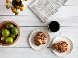 Overhead view of a light wooden table with green apples, baked muffins on white plates, a cup of coffee, and a newspaper.