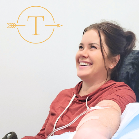 Smiling woman receives IV drip with "T" logo in comfortable setting.