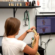 Woman holds anatomical spine model beside monitor with body diagrams.