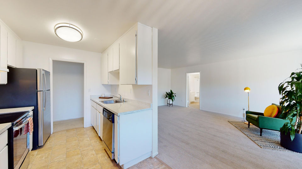 A panoramic view of an open-concept living space, showing a kitchen with white cabinets, stainless steel appliances, and light-colored tile flooring on the left, transitioning into a living room with light beige carpeting, a dark green armchair with an orange pillow, a yellow floor lamp, and a large green plant. A doorway to the bathroom is visible on the left.
