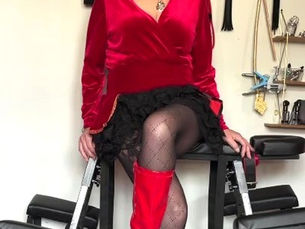 Dominant woman in velvet and PVC outfit seated in a controlled setting.