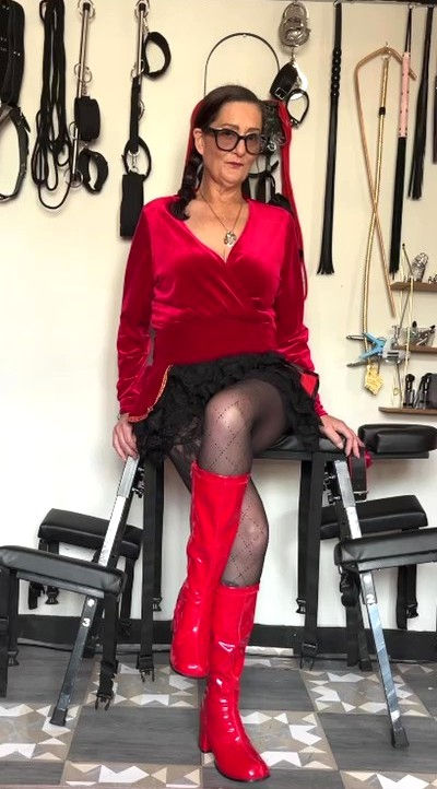 Dominant woman in velvet and PVC outfit seated in a controlled setting.