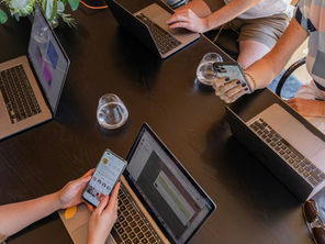 Four people work at a black table with laptops, two holding smartphones, and three glasses of water visible among the devices.