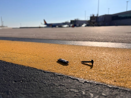 Screw and small object on a runway with a yellow line in focus; blurred plane and terminal in the sunny background.