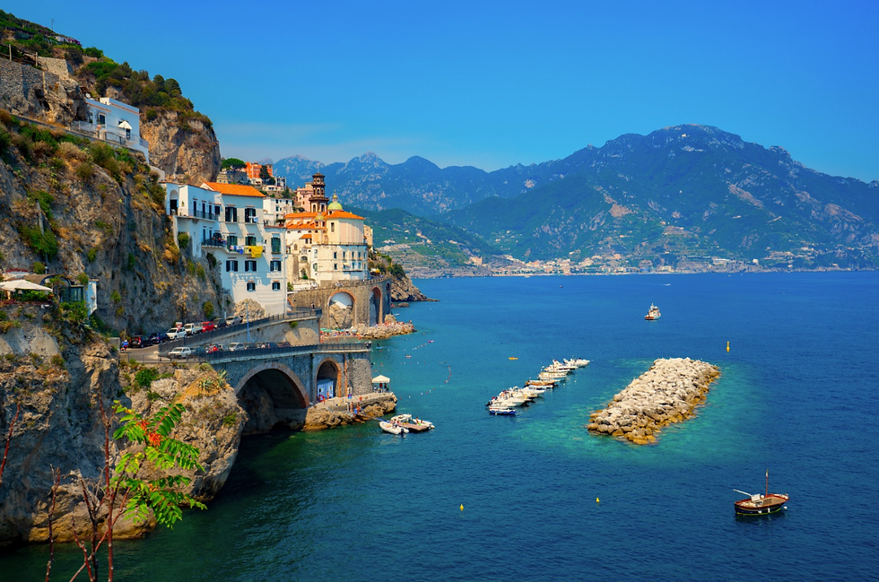Cliffside village with colourful buildings overlooking a blue sea. Boats docked on a rocky pier. Mountains in the background, sunny day.