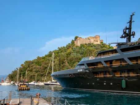 Luxury yacht named "MAIN" docked in a scenic harbour with sailboats. Green hills and an old fort in the background under a clear blue sky.