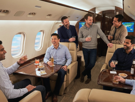 Seven men socialize with drinks in a luxurious private jet. Smiling, seated around tables with snacks. Bright, relaxed atmosphere.