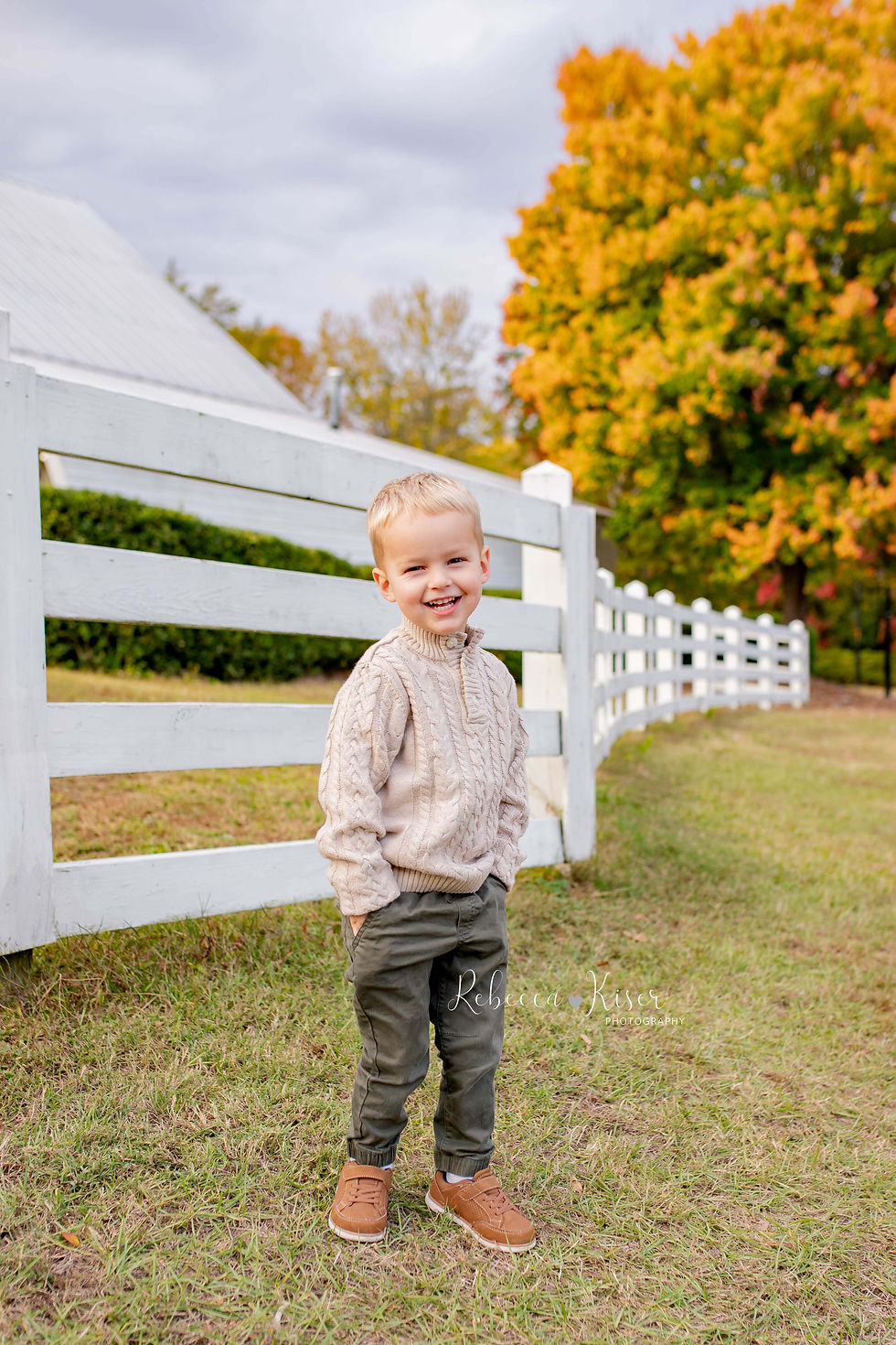Young child in a beige sweater and green pants stands smiling for his fall family portraits, by a white fence. Autumn trees in the background. Warm, cheerful mood.