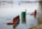 Flooded riverside park with submerged wooden benches and a green trash can. Water covers the ground, creating a calm, somber scene.