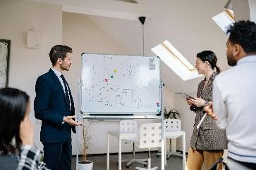 Business meeting in a bright room. A man in a suit presents at a whiteboard with diagrams. Attendees listen, holding notebooks.