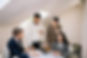 Four colleagues discuss documents at a table in an office. Two hold coffee cups. Bright lighting from skylights creates a focused mood.
