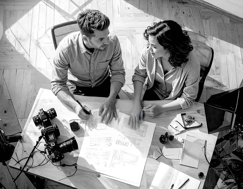 Two people in shirts discuss blueprints at a wooden desk with cameras and sketches, in a bright room with a creative and collaborative mood.