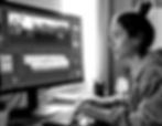 A woman edits a video on a computer in a room, focused expression, grayscale image. Linear patterns on the screen, natural light behind.