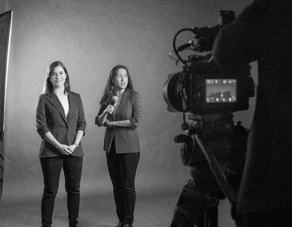 Two women in suits are being filmed in a studio. One holds a microphone. Black and white setting, focused lighting. They appear calm.