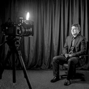 Man in a suit sits on a chair, smiling for a camera in a studio with dark curtains and bright spotlight. Black and white image.