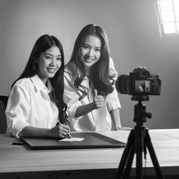 Two women in white shirts smile at a camera on a tripod, with one giving a thumbs-up, in a studio with bright lighting. Black and white photo.