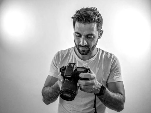 Man in white shirt examines camera, focused expression. Neutral background with spotlights. Black and white image.