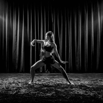 Dancer in athletic wear striking a pose on a dimly lit stage with velvet curtains; dramatic lighting creates an intense, focused mood.