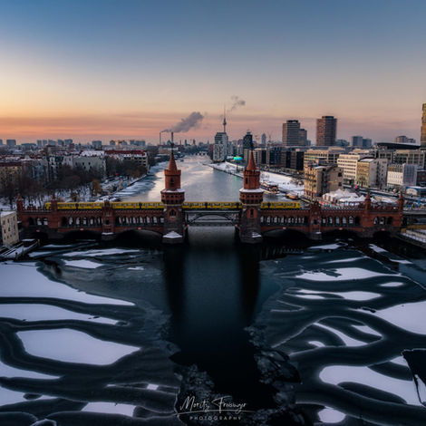 Seltene Winteraufnahme der vereisten Spree in Berlin mit der Oberbaumbrücke im Fokus. Eine gelbe U-Bahn der Linie U1 überquert die historische Backsteinbrücke, während im Hintergrund die Berliner Skyline mit dem Fernsehturm unter einem klaren Winterhimmel sichtbar ist. Hochwertige Cityscape-Fotografie im Schnee von Moritz Freisinger, erhältlich als Fine Art Print
