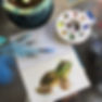 A top-down photograph of a watercolor sketchbook on a glass table surrounded by watercolor pens and a painting palette. The sketchbook features a watercolor painting of a frog.