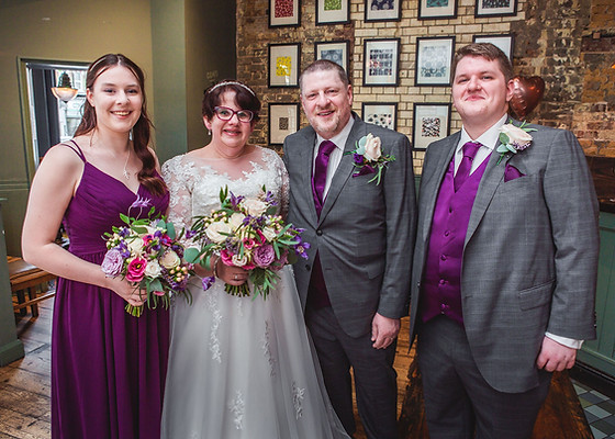 Married couple standing with their best man and bridesmaid in a grape-coloured dress, carrying pink, purple and ivory wedding bouquets.