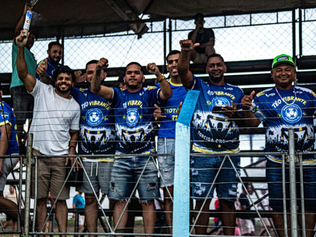 Torcedores com camisas azul e amarelo "Teo Vibrante" celebram em arquibancada de estádio. Emoção e entusiasmo no ar.