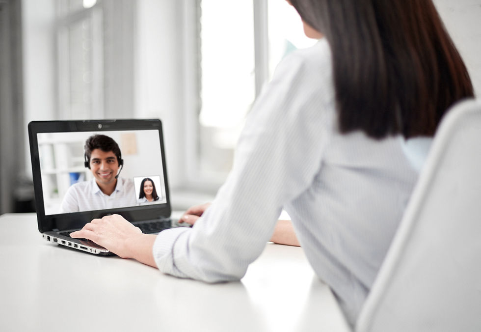 Person in white shirt on video call using laptop, showing two people on screen. Bright office setting with natural light from windows.